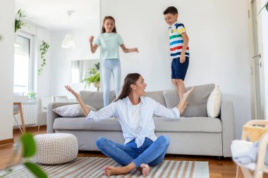 Küçük çocuklar kanepede zıplayıp eğlenirken ve gürültü yaparken genç anne yerde oturup dizüstü bilgisayarla ve belgelerle çalışıyor.