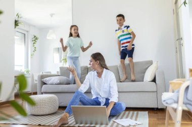 Küçük çocuklar kanepede zıplayıp eğlenirken ve gürültü yaparken genç anne yerde oturup dizüstü bilgisayarla ve belgelerle çalışıyor.