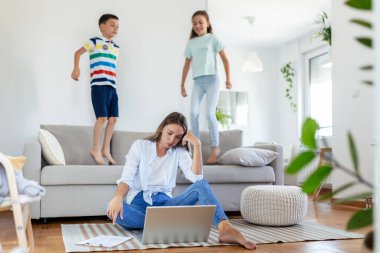 Küçük çocuklar kanepede zıplayıp eğlenirken ve gürültü yaparken genç anne yerde oturup dizüstü bilgisayarla ve belgelerle çalışıyor.
