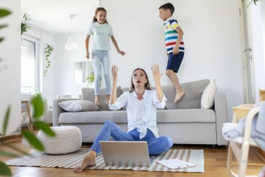 Küçük çocuklar kanepede zıplayıp eğlenirken ve gürültü yaparken genç anne yerde oturup dizüstü bilgisayarla ve belgelerle çalışıyor.