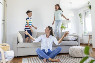 Küçük çocuklar kanepede zıplayıp eğlenirken ve gürültü yaparken genç anne yerde oturup dizüstü bilgisayarla ve belgelerle çalışıyor.