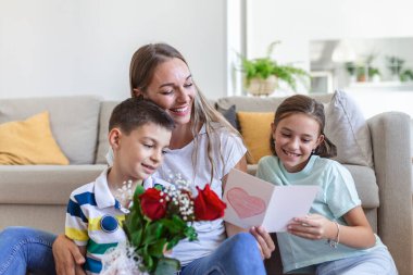 Elinde gül buketi olan genç bir anne gülüyor, oğluna sarılıyor ve elinde bir kart ve güllerle korkunç bir kız evde, mutfakta tatil kutlamalarında annemi tebrik ediyor. Anneler Günü