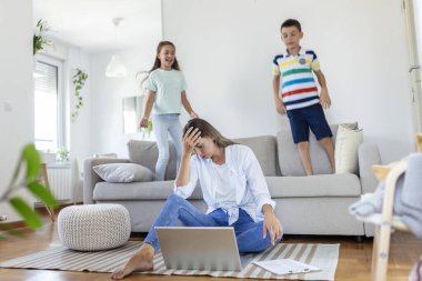Küçük çocuklar kanepede zıplayıp eğlenirken ve gürültü yaparken genç anne yerde oturup dizüstü bilgisayarla ve belgelerle çalışıyor.