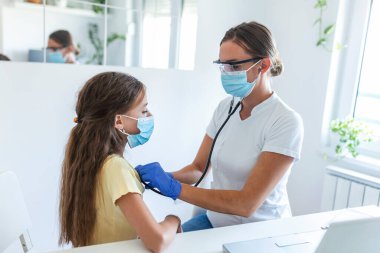 Sevimli kız doktorunun ofisinde oturuyor. Doktoru göğsünü dinlemek için steteskop kullanıyor. Hem o hem de doktoru mikropların yayılmasını önlemek için maske takıyor..