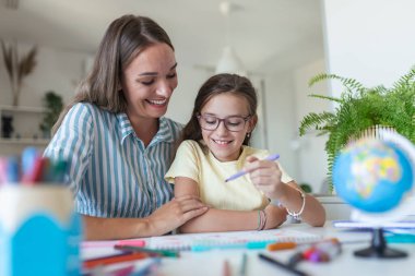 Anne, kızına evde ders çalışırken yardım ediyor. Güzel, gülümseyen bir kadın, sevimli kızına evde ödevlerini yapmasına yardım ediyor.