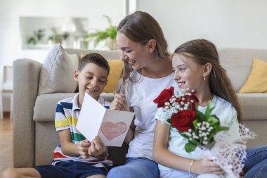 Elinde gül buketi olan genç bir anne gülüyor, oğluna sarılıyor ve elinde kart ve güllerle korkunç bir kız evde tatil kutlamalarında annesini tebrik ediyor. Anneler Günü