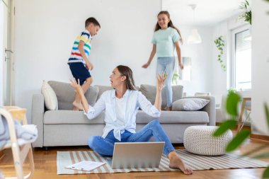 Küçük çocuklar kanepede zıplayıp eğlenirken ve gürültü yaparken genç anne yerde oturup dizüstü bilgisayarla ve belgelerle çalışıyor.