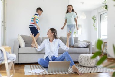Küçük çocuklar kanepede zıplayıp eğlenirken ve gürültü yaparken genç anne yerde oturup dizüstü bilgisayarla ve belgelerle çalışıyor.