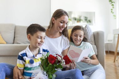 Elinde gül buketi olan genç bir anne gülüyor, oğluna sarılıyor ve elinde bir kart ve güllerle korkunç bir kız evde, mutfakta tatil kutlamalarında annemi tebrik ediyor. Anneler Günü