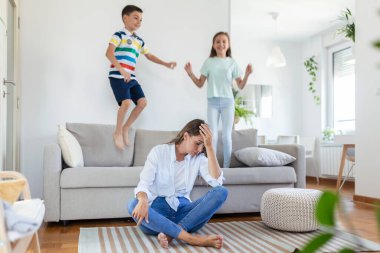 Küçük çocuklar kanepede zıplayıp eğlenirken ve gürültü yaparken genç anne yerde oturup dizüstü bilgisayarla ve belgelerle çalışıyor.