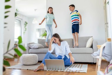 Küçük çocuklar kanepede zıplayıp eğlenirken ve gürültü yaparken genç anne yerde oturup dizüstü bilgisayarla ve belgelerle çalışıyor.