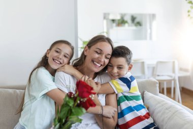 Elinde gül buketi olan genç bir anne gülüyor, oğluna sarılıyor ve elinde bir kartla korkunç bir kız, mutfakta tatilde annesini tebrik ediyor.