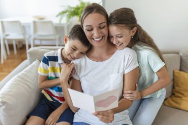 Mutlu küçük anne ve çocuklar kanepede oturup dileklerini okurken kucaklaşıyorlar ve evde sunulan kartpostalda tatille kutluyorlar.