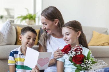 Elinde gül buketi olan genç bir anne gülüyor, oğluna sarılıyor ve elinde kart ve güllerle korkunç bir kız evde tatil kutlamalarında annesini tebrik ediyor. Anneler Günü
