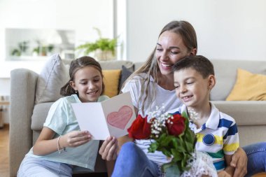 Elinde gül buketi olan genç bir anne gülüyor, oğluna sarılıyor ve elinde bir kart ve güllerle korkunç bir kız evde, mutfakta tatil kutlamalarında annemi tebrik ediyor. Anneler Günü