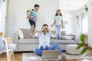 Küçük çocuklar kanepede zıplayıp eğlenirken ve gürültü yaparken genç anne yerde oturup dizüstü bilgisayarla ve belgelerle çalışıyor.