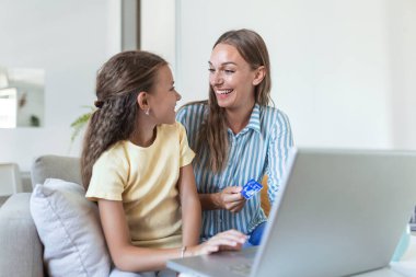Düşük açılı mutlu küçük kız sevinirken günlük kıyafetlerle gülümseyen anne ekrana odaklanıyor ve hafif modern apartmanda online alışveriş için dizüstü bilgisayar kullanıyor.