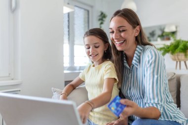 Düşük açılı mutlu küçük kız sevinirken günlük kıyafetlerle gülümseyen anne ekrana odaklanıyor ve hafif modern apartmanda online alışveriş için dizüstü bilgisayar kullanıyor.
