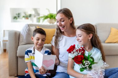 Elinde gül buketi olan genç bir anne gülüyor, oğluna sarılıyor ve elinde kart ve güllerle korkunç bir kız evde tatil kutlamalarında annesini tebrik ediyor. Anneler Günü