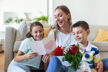 Elinde gül buketi olan genç bir anne gülüyor, oğluna sarılıyor ve elinde bir kart ve güllerle korkunç bir kız evde, mutfakta tatil kutlamalarında annemi tebrik ediyor. Anneler Günü