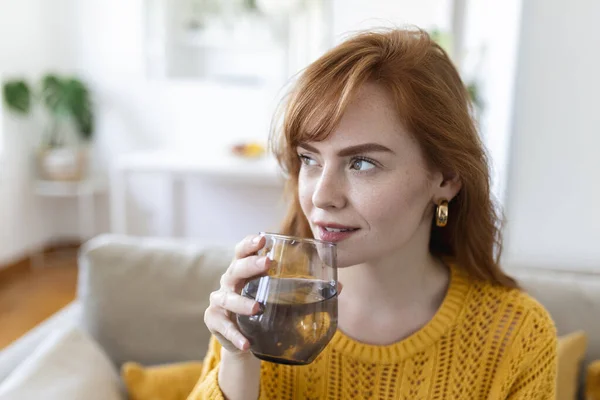 Happy young woman smiling while holding a glass of water at home. Woman on living room sofa Relaxed and Smiling While Drinking glass of water. Health benefits of drinking enough water