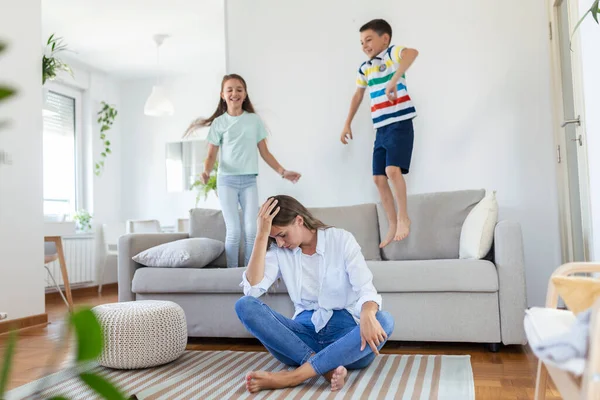 Küçük çocuklar kanepede zıplayıp eğlenirken ve gürültü yaparken genç anne yerde oturup dizüstü bilgisayarla ve belgelerle çalışıyor.