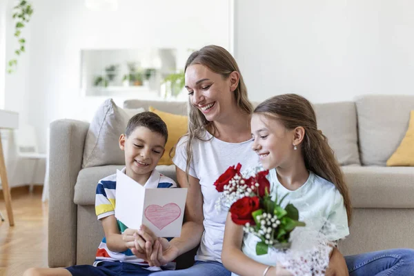 Young mother with a bouquet of roses laughs, hugging her son, and ?heerful girl with a card and roses congratulates mom during holiday celebration at home. mothers day