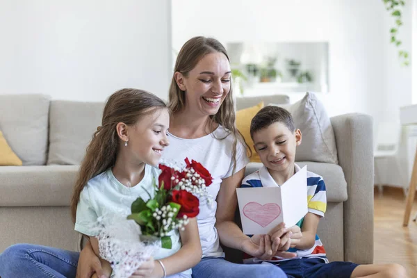 Elinde gül buketi olan genç bir anne gülüyor, oğluna sarılıyor ve elinde kart ve güllerle korkunç bir kız evde tatil kutlamalarında annesini tebrik ediyor. Anneler Günü