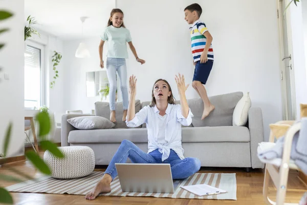 Küçük çocuklar kanepede zıplayıp eğlenirken ve gürültü yaparken genç anne yerde oturup dizüstü bilgisayarla ve belgelerle çalışıyor.