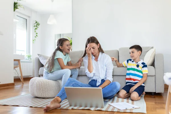 Small mischievous kids noising and distracting mother freelancer trying to concentrate on laptop and squeezing head working at table in light living room