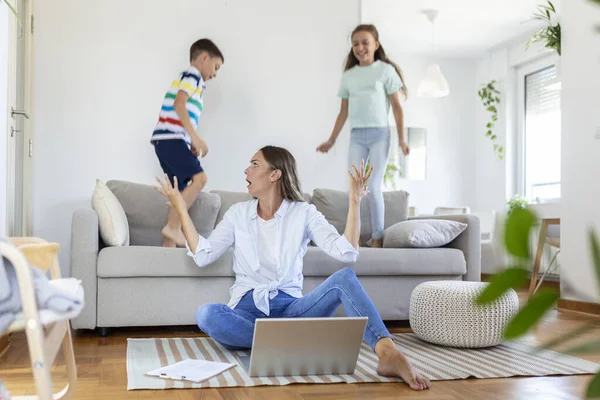 Küçük çocuklar kanepede zıplayıp eğlenirken ve gürültü yaparken genç anne yerde oturup dizüstü bilgisayarla ve belgelerle çalışıyor.