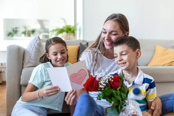 Elinde gül buketi olan genç bir anne gülüyor, oğluna sarılıyor ve elinde bir kart ve güllerle korkunç bir kız evde, mutfakta tatil kutlamalarında annemi tebrik ediyor. Anneler Günü