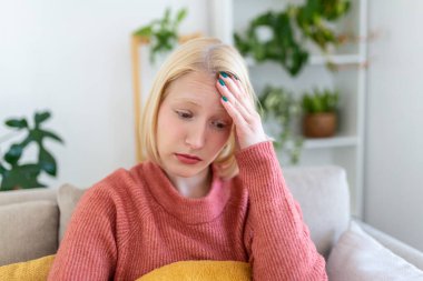 Portrait of a young blond woman sitting on the couch at home with a headache and migraine . Beautiful woman suffering from chronic daily headaches. Sad woman holding her head because sinus pain