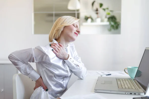 Portrait of young stressed woman sitting at home office desk in front of laptop, touching aching neck with pained expression, suffering from neck pain after working on laptop