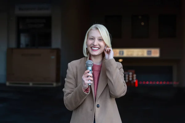 Cropped portrait of professional female reporter at work. Young woman standing on the street with a microphone in hand and smiling at camera. Horizontal shot. Selective focus on woman