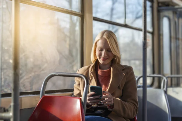 Modern smiling cute woman sitting in a bus . Smiling and looking away. Bus journey. Woman using her cell phone on bus. Tramway. Sms, message. Woman with phone at the public transport