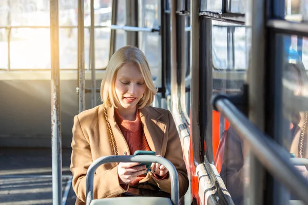 Modern smiling cute woman sitting in a bus . Smiling and looking away. Bus journey. Woman using her cell phone on bus. Tramway. Sms, message. Woman with phone at the public transport