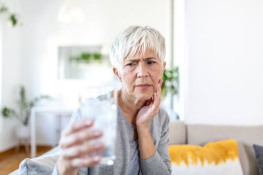 Hassas dişleri olan ve elinde buzlu soğuk su bardağı tutan yaşlı bir kadın. Sağlık sigortası konsepti. Olgun bir kadın soğuk içecek içiyor, bir bardak dolusu buz ve diş ağrısı çekiyor, acı çekiyor.