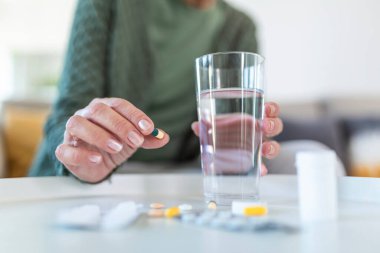 Sağlık sigortası konsepti. Yaşlı bir kadın vitamin hapı içerken, elinde su olan bardağı tutarken, ahşap masanın arkasında tek başına otururken görüntülenmiş.
