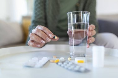 Sağlık sigortası konsepti. Yaşlı bir kadın vitamin hapı içerken, elinde su olan bardağı tutarken, ahşap masanın arkasında tek başına otururken görüntülenmiş.
