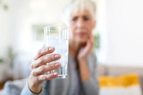 Senior woman with sensitive teeth and hand holding glass of cold water with ice. Healthcare concept. Mature woman drinking cold drink, glass full of ice cubes and feels toothache, pain