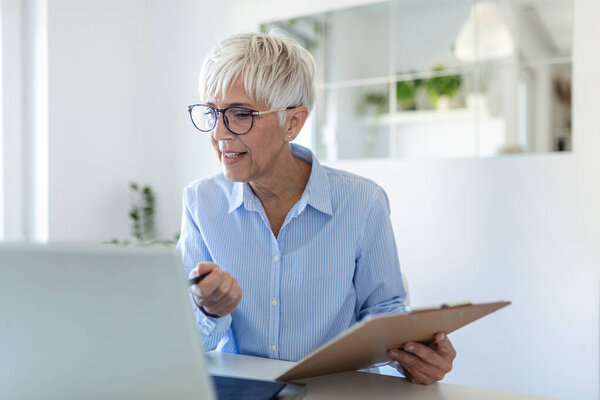 Elderly woman working on laptop computer, smiling. Woman Working From Home On Laptop In Modern Apartment. Trendy woman working on laptop from home