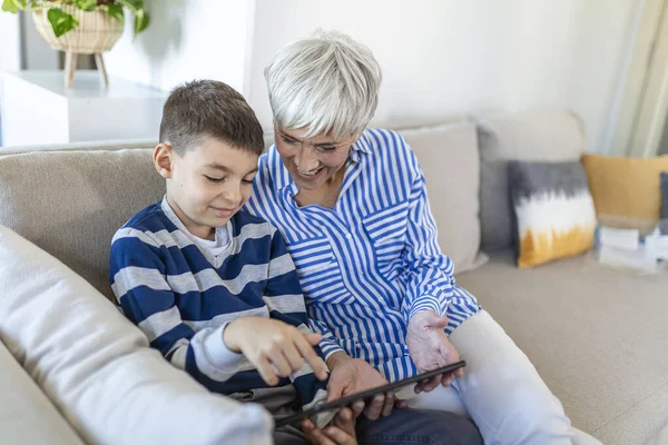 Happy grandmother and grandson using tablet together, sitting on cozy sofa at home, browsing tablet device apps, grandma with grandchild playing game, looking at screen, having fun