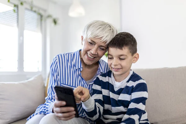 Happy grandmother and grandson using tablet together, sitting on cozy sofa at home, browsing tablet device apps, grandma with grandchild playing game, looking at screen, having fun