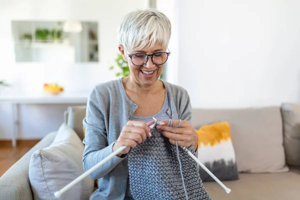Elderly woman in glasses sit on couch at home smile holding knitting needles and yarn knits clothes for loved ones, favorite activity and pastime, retired tranquil carefree life concept