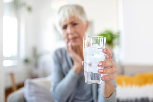 Senior woman with sensitive teeth and hand holding glass of cold water with ice. Healthcare concept. Mature woman drinking cold drink, glass full of ice cubes and feels toothache, pain