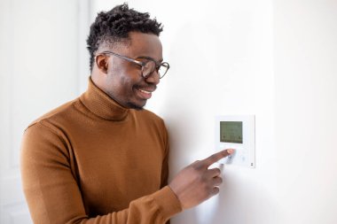 Gülümseyen Afrikalı Amerikalı adam modern akıllı ev sistemini kullanıyor, duvarda kontrolör var, pozitif genç adam termostat ısısını değiştiriyor ya da apartmandaki güvenlik alarmını çalıştırıyor.