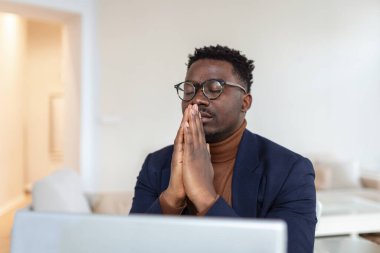 African guy sit at desk with clasped palms closed eyes thinking search problem solution, collect thoughts together, organize his emotional state before important meeting. Prayer hands sign of ask help
