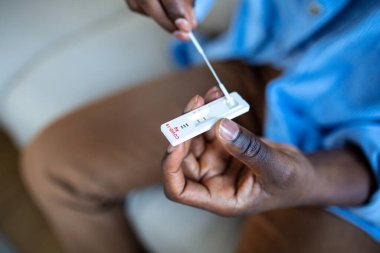 Genç Afrikalı Amerikalı adam evde antijen kiti ile COVID-19 testi yaptırıyor. Coronavirus enfeksiyonunu kontrol etmek için burun sopasıyla giriyoruz. Karantina, salgın hastalık.