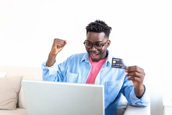 Internet payment. Happy cheerful man looking at the laptop screen while making an Internet payment. Happy young man with credit card and laptop sitting on sofa at home. Online payment